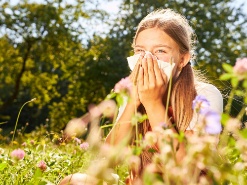 A young girl blows her nose into a tissue while she sits among a field of wildflowers. Get tested for allergies at The Allergy and Asthma Group allergy fair.